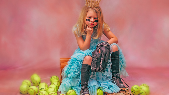 A young girl sits posed around softballs in a blue dress.