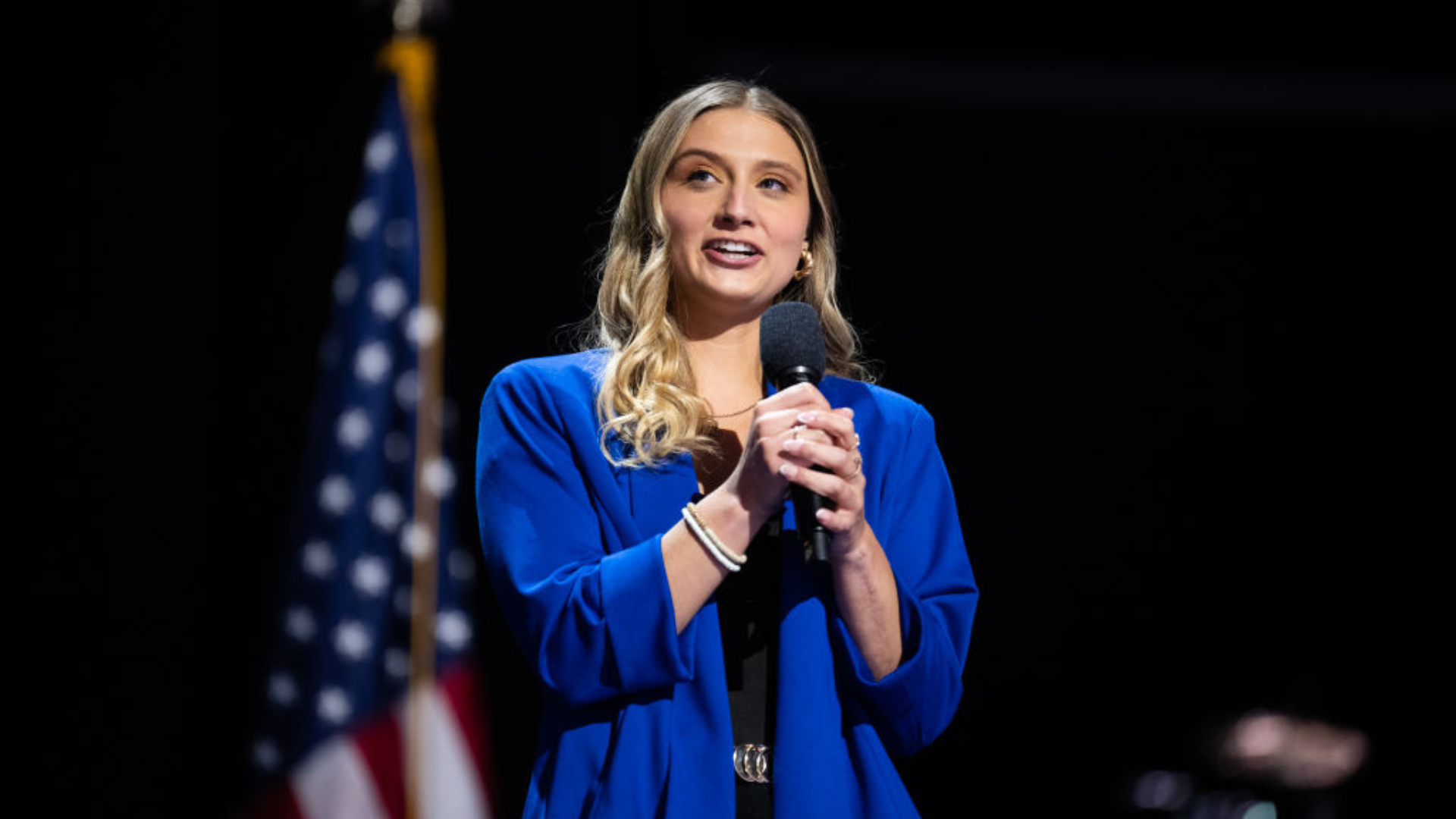 Hadley Duvall in a blue blazer holding a microphone in front of an American flag