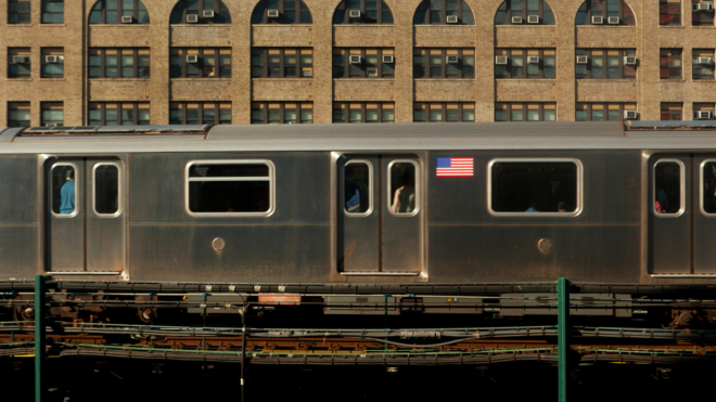a New York City subway car on elevated track against a wall with many windows