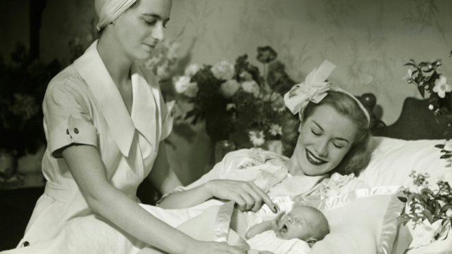 black and white photo of mother laying in a hospital bed holding a newborn while a nurse checks on them