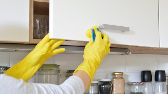 Woman in yellow gloves cleans kitchen cabinets.