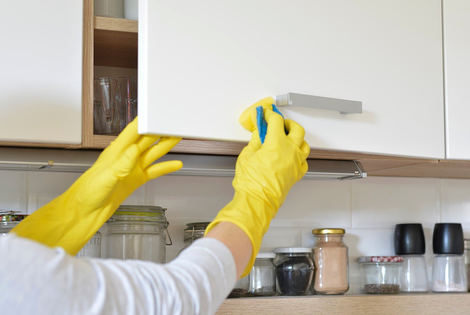 Woman in yellow gloves cleans kitchen cabinets.