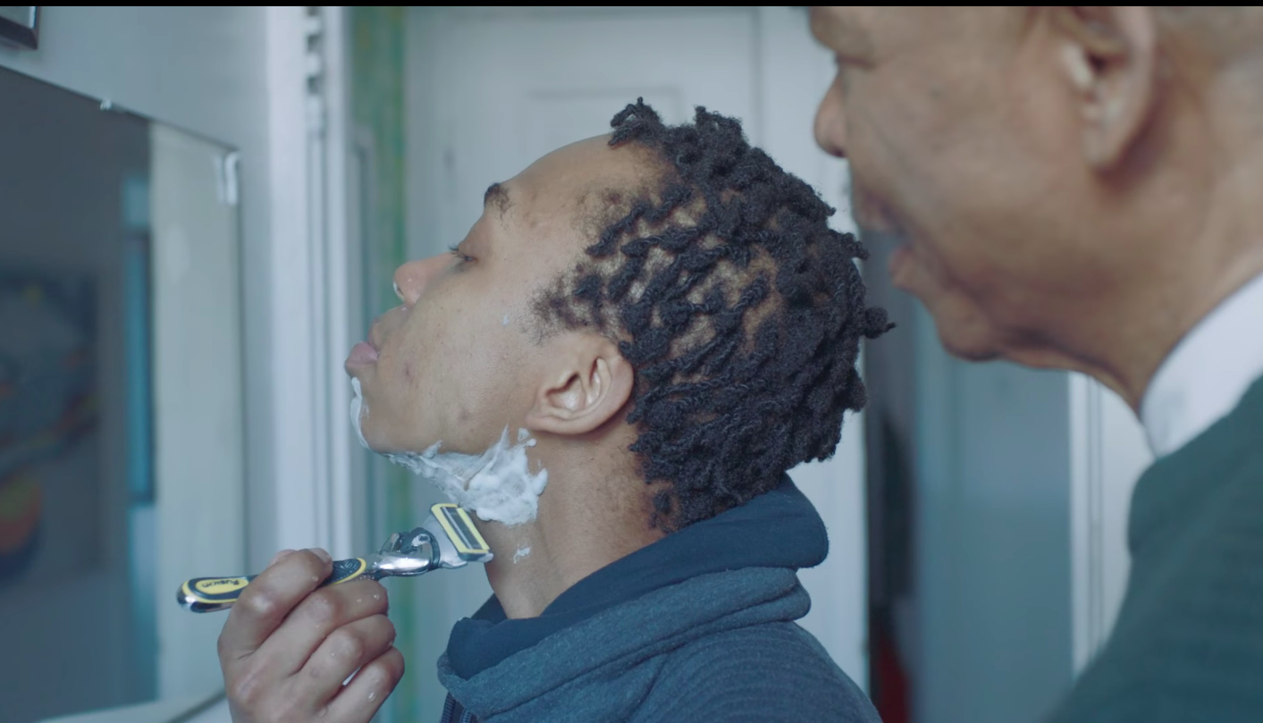 A young trans man attempts his first shave as his dad looks on.