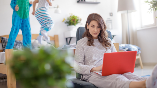 mom wearing pjs working while kids in pjs are jumping on the bed