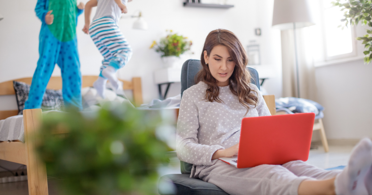 mom wearing pjs working while kids in pjs are jumping on the bed