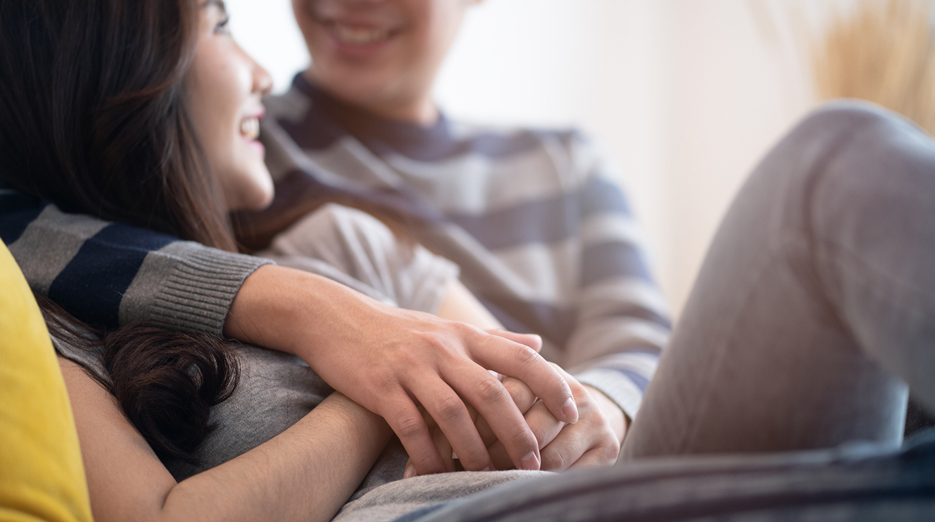 Lovely Asian couple sitting on bed in bedroom talking together with happiness. Boy hugging girl and holding hand, be hand in hand. Crop shot focus on hands. Man looking at woman eyes and smile face.