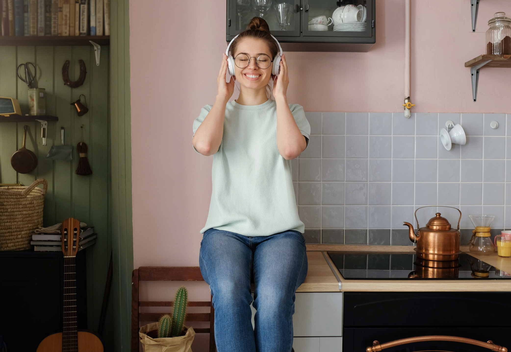 Indoor photo of young positive European Caucasian lady spending free time at home sitting on kitchen table listening to favorite music through headphones, closed her eyes in delight and relaxation