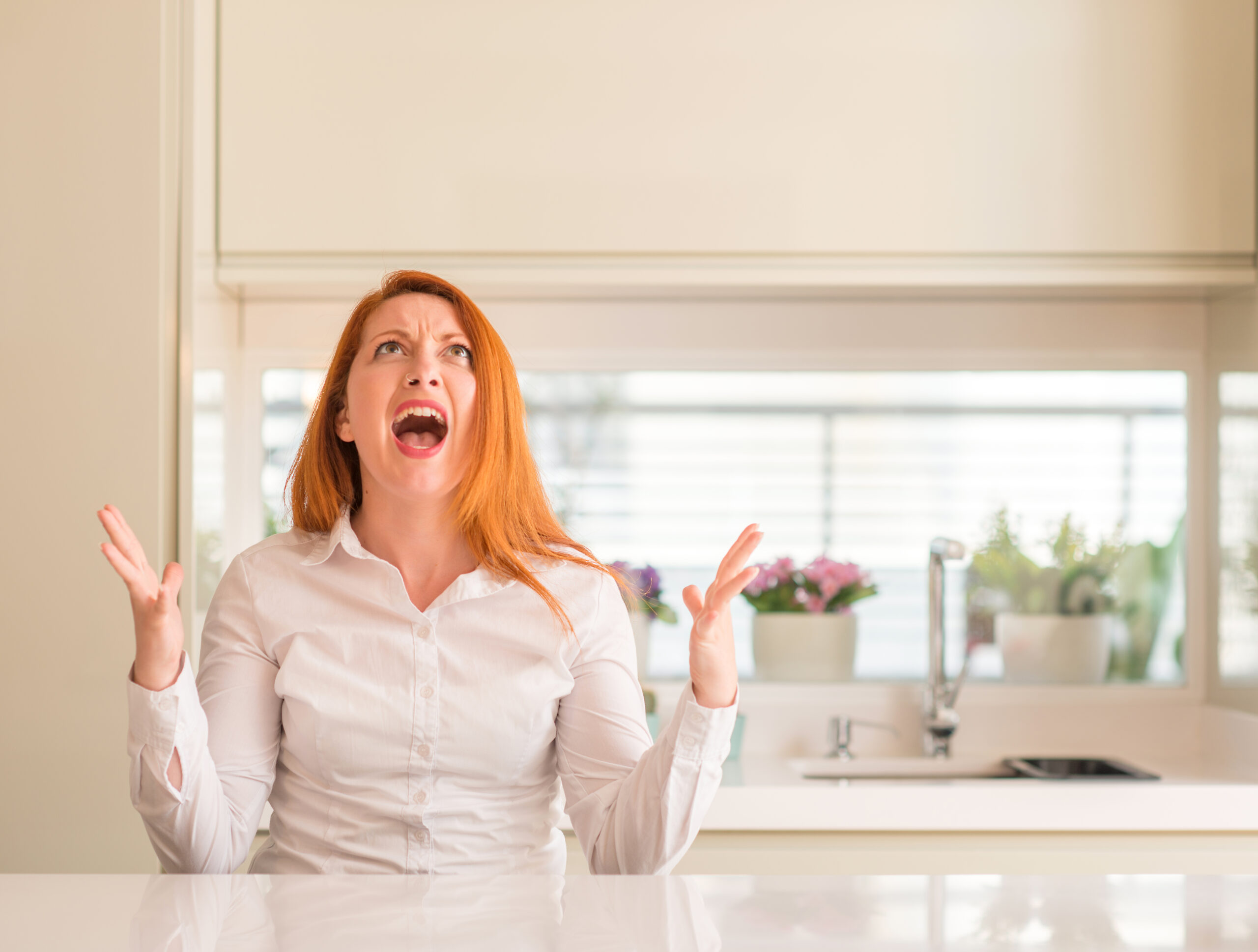 Redhead woman at kitchen crazy and mad shouting and yelling with aggressive expression and arms raised. Frustration concept.