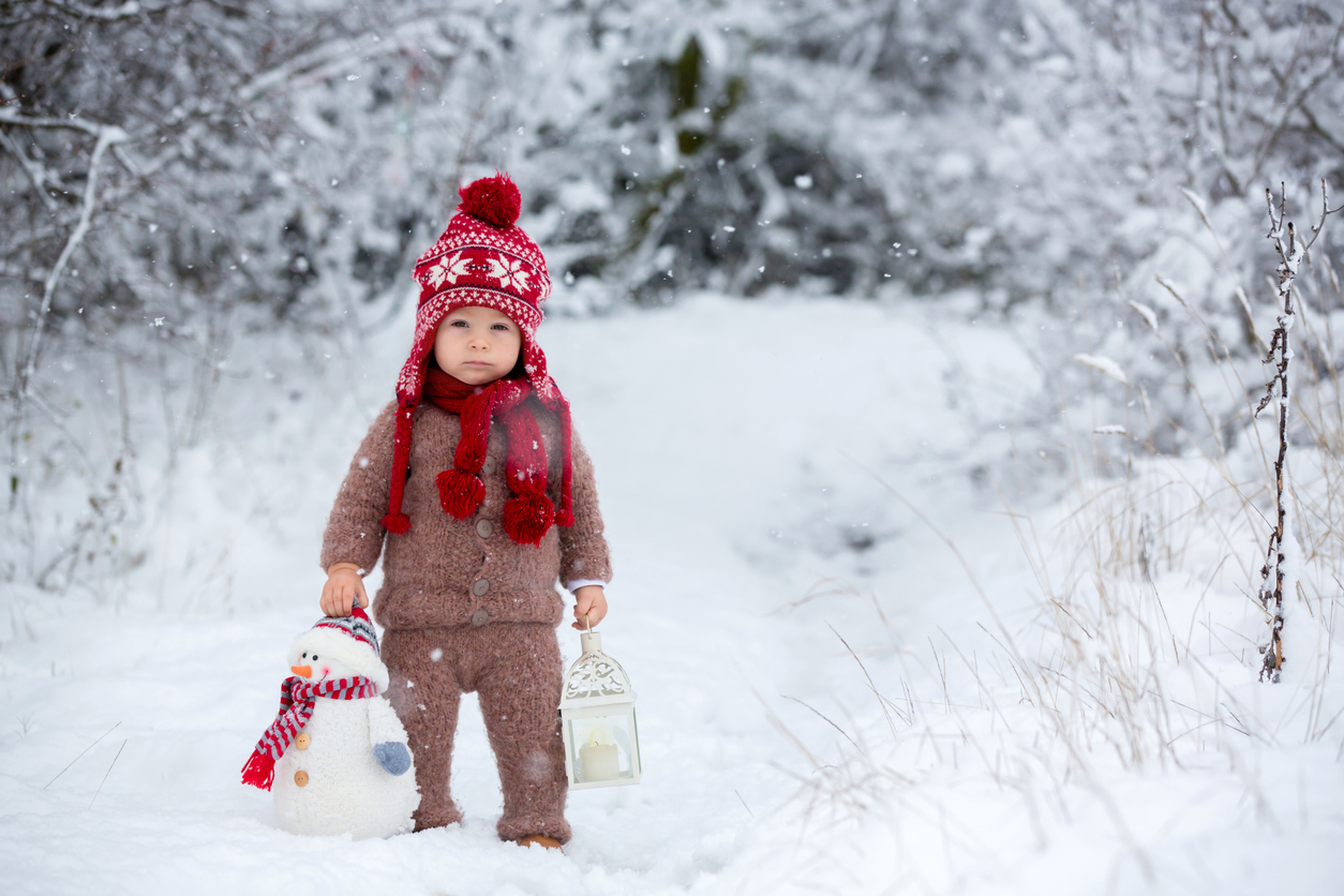 Portrait of a cute toddler baby dressed in a brown hand knitted jacket, pants, red hat and scarf, holding teddy and lantern, walks through the snowy park enjoying first snow blowing
