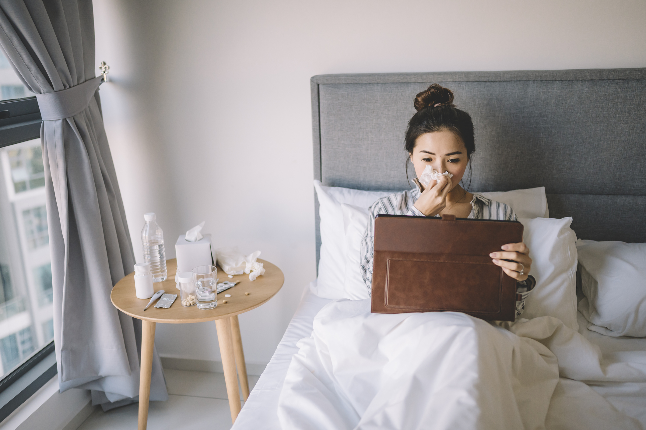 an asian chinese femela reading digital tablet while holding her nose with tissue on her bed with her pajamas