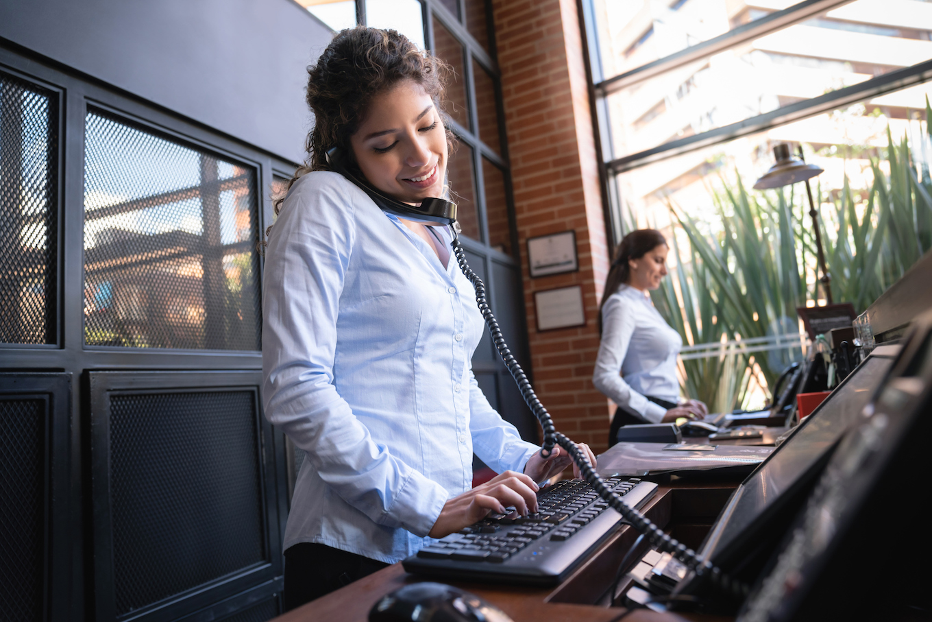 Beautiful hotel receptionist on a phone call looking at the bookings on system smiling