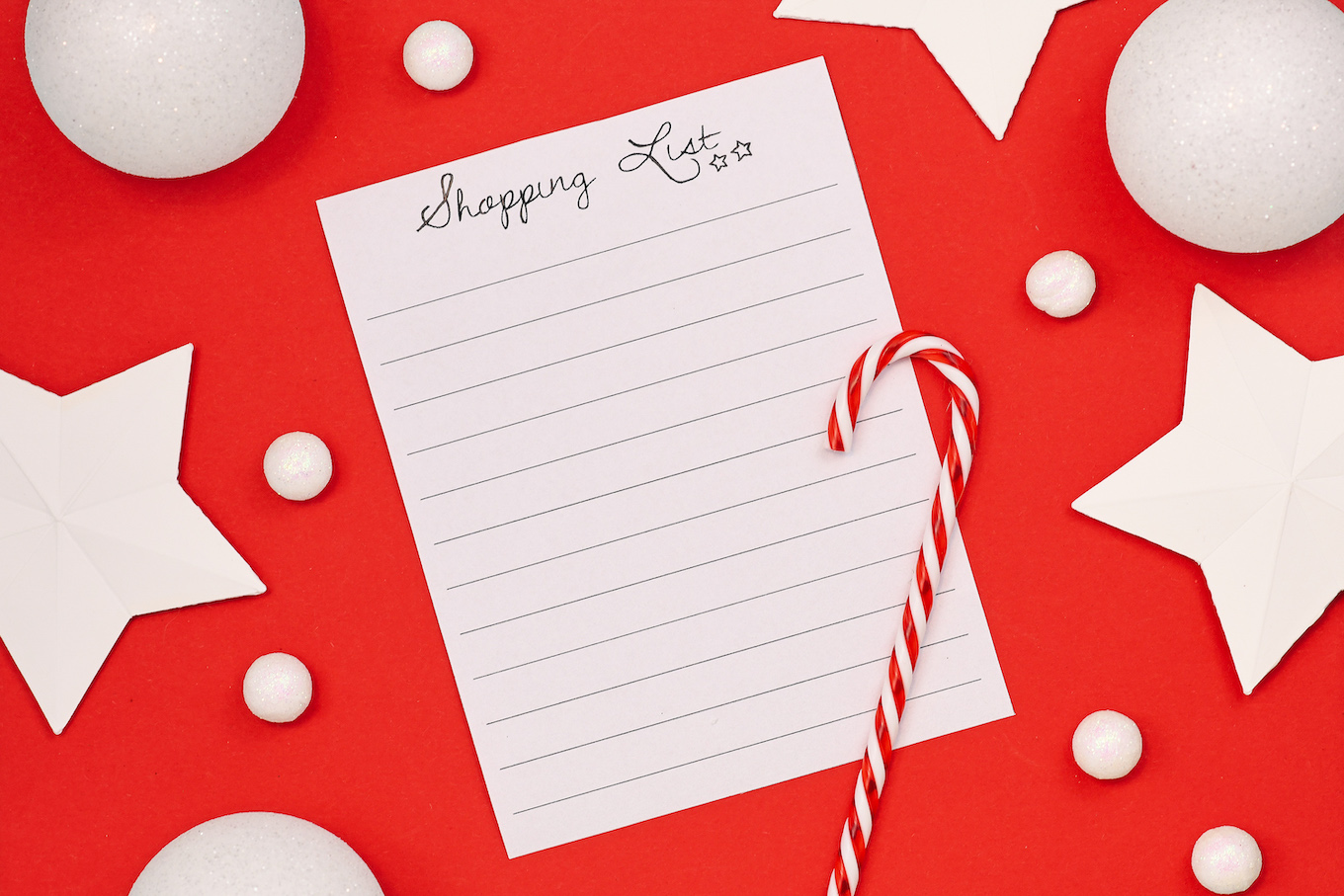 Concept for Christmas shopping with top view flat lay with empty shopping check list and seasonal decorations like candy cane, snow and stars on bright red background
