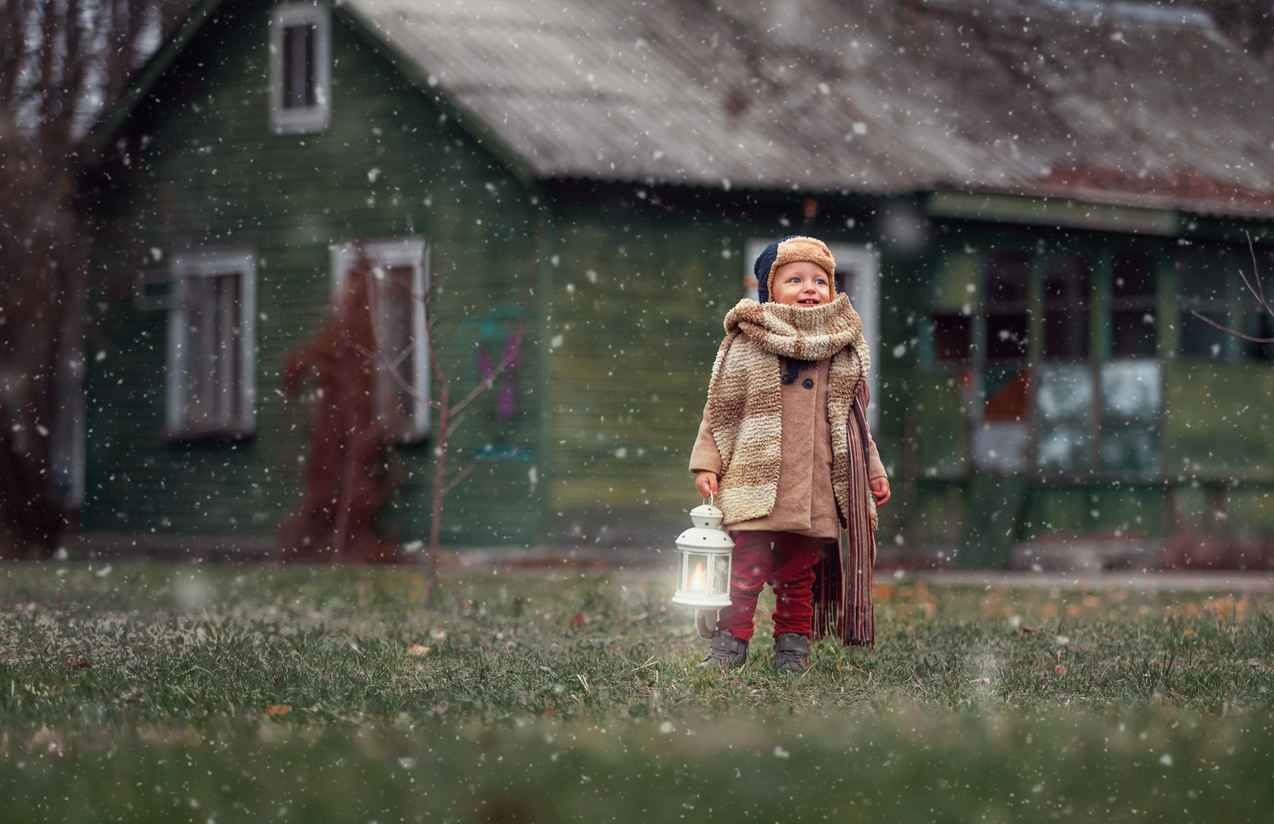Sirst snow. little kid having enjoying countryside. Baby boy playing outdoors with vintage hand lamp, winter time old wooden house on background.