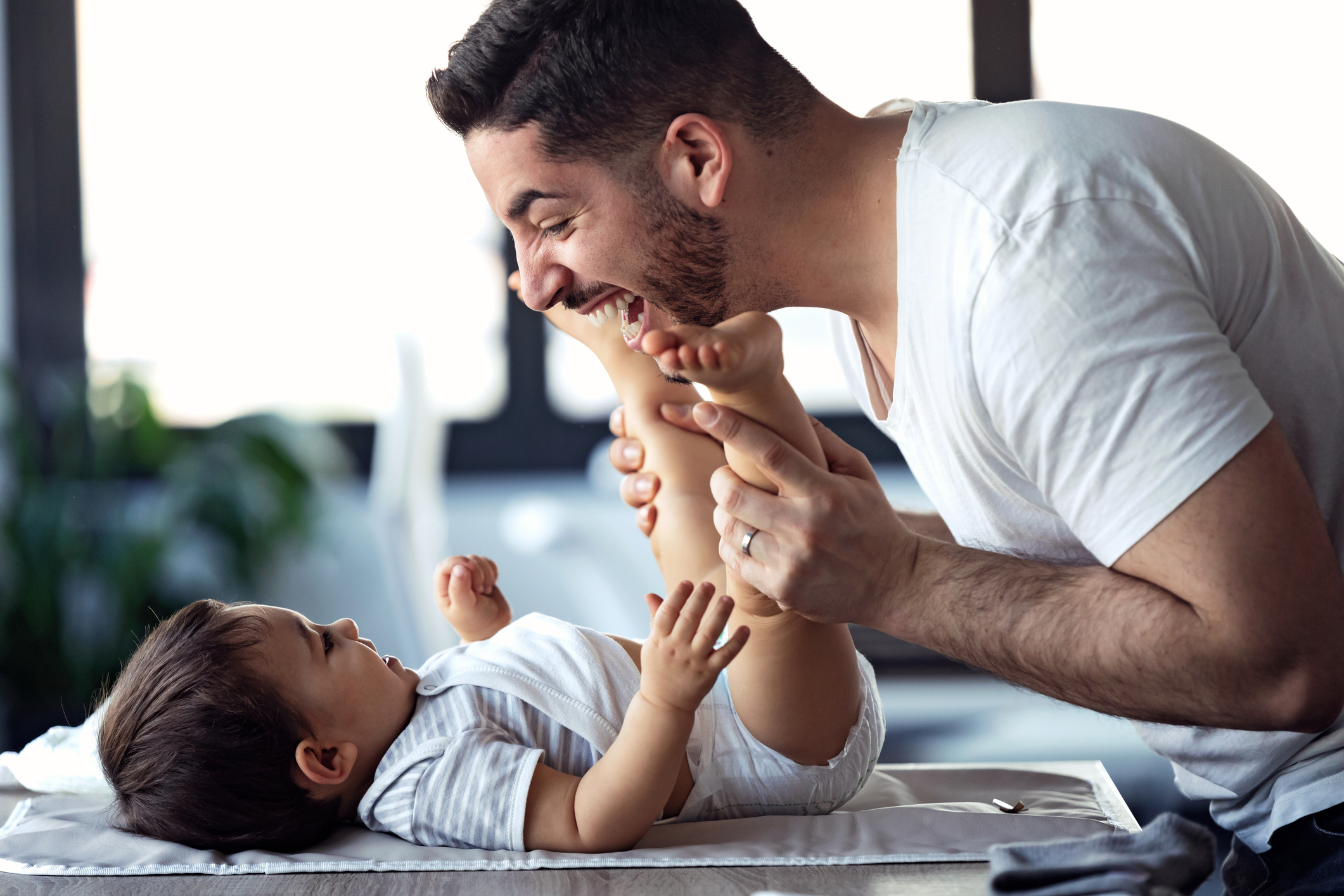 Smiling young father has fun with little baby while changing his nappy at home.