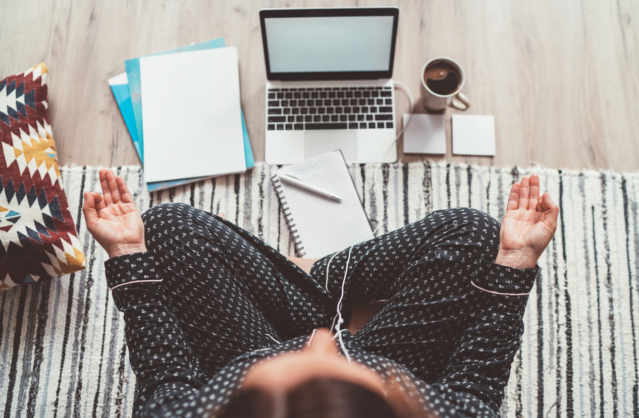 Businesswoman dressed in pajamas, listening earphones meditating with morning coffee with Gyan Mudra palm on floor office with laptop, papers top view shot. Distance work in quarantine time concept.