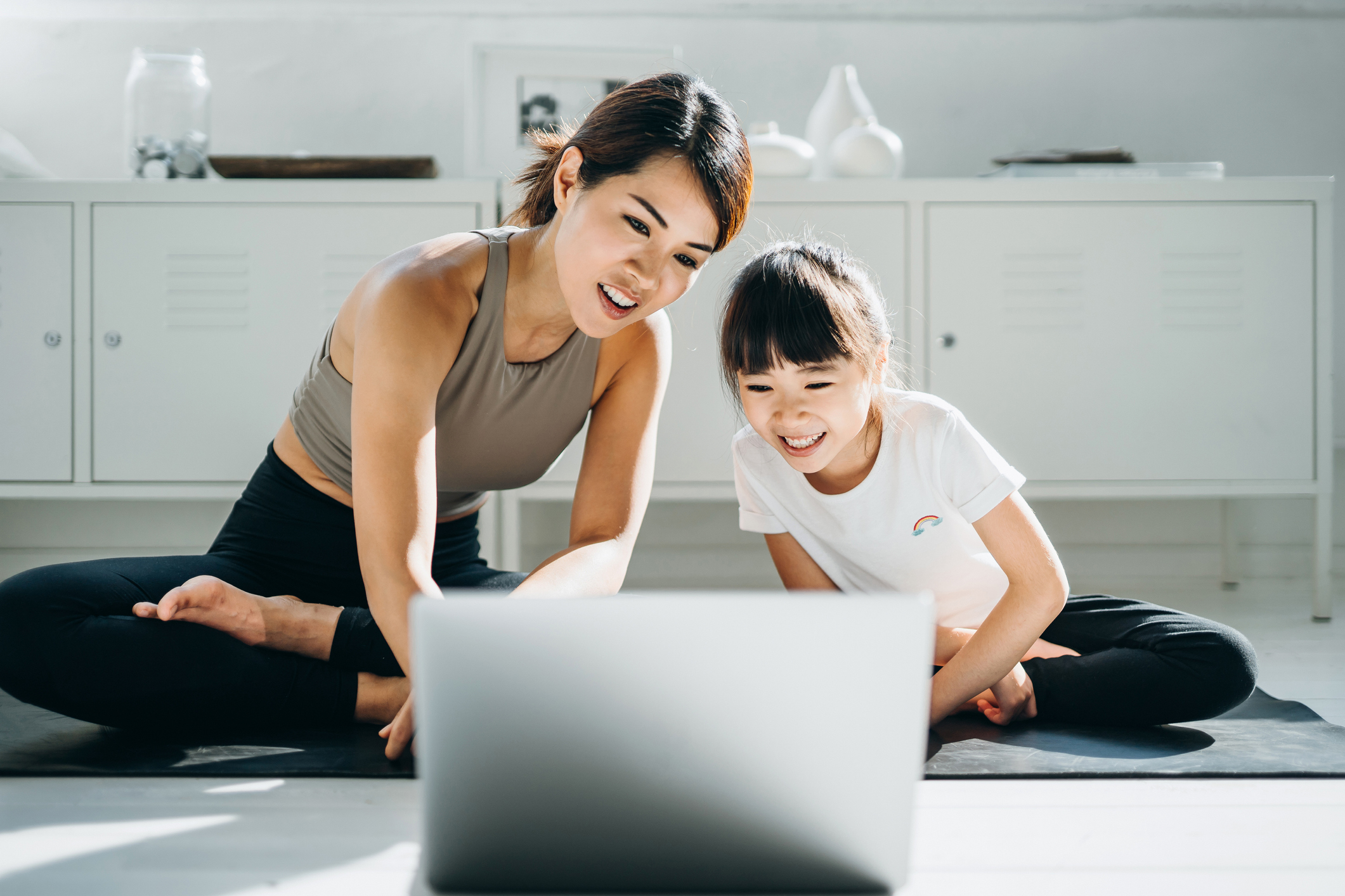 Joyful young Asian mother and little daughter doing online yoga class together with a laptop at home