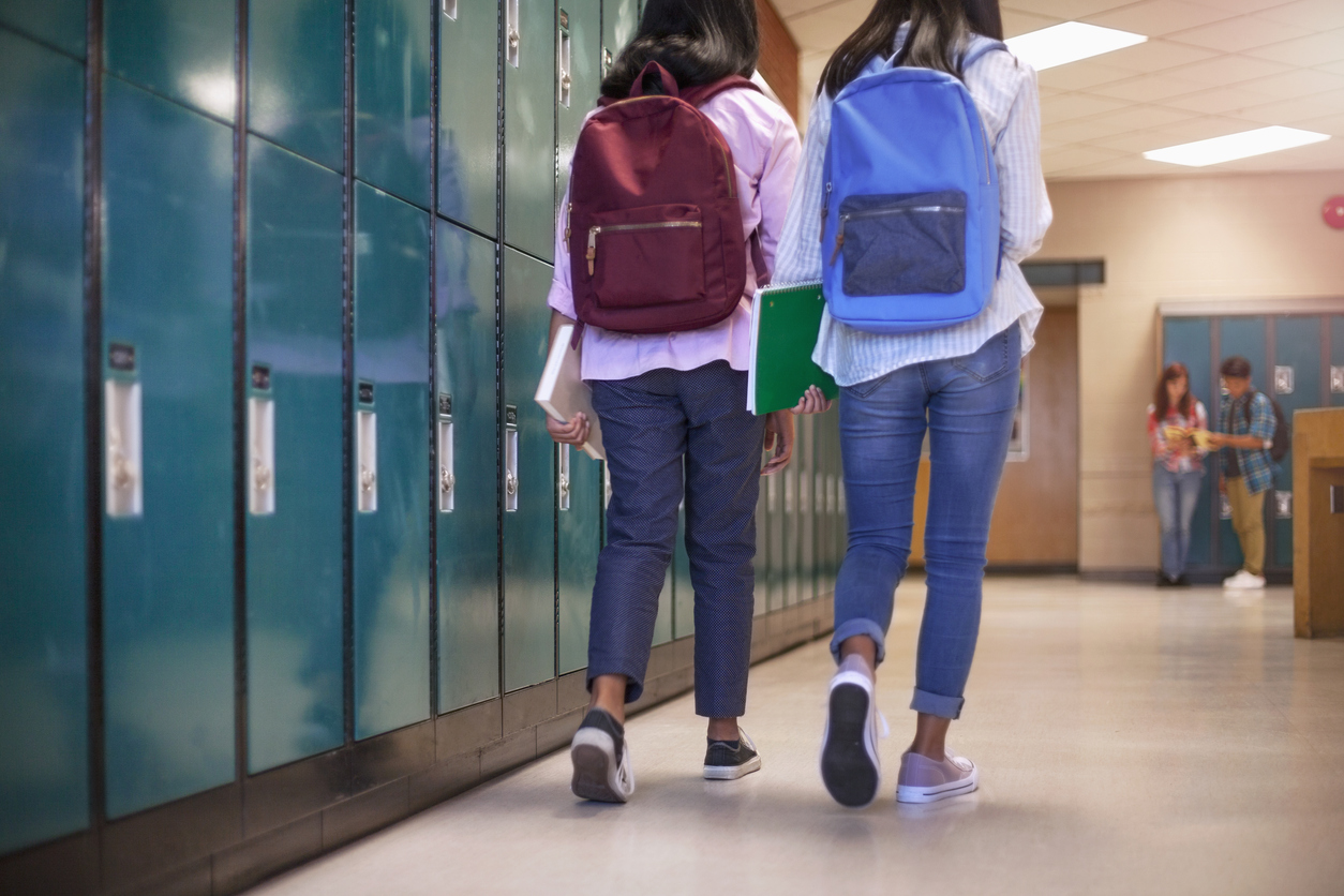 Female classmate friends with backpacks walking by lockers in school