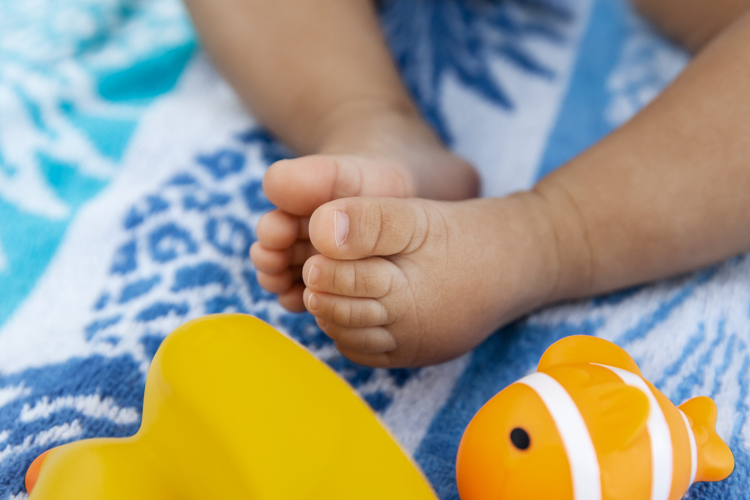 Little infant legs of a baby girl sitting on a beach towel and playing with rubber toys at summer time