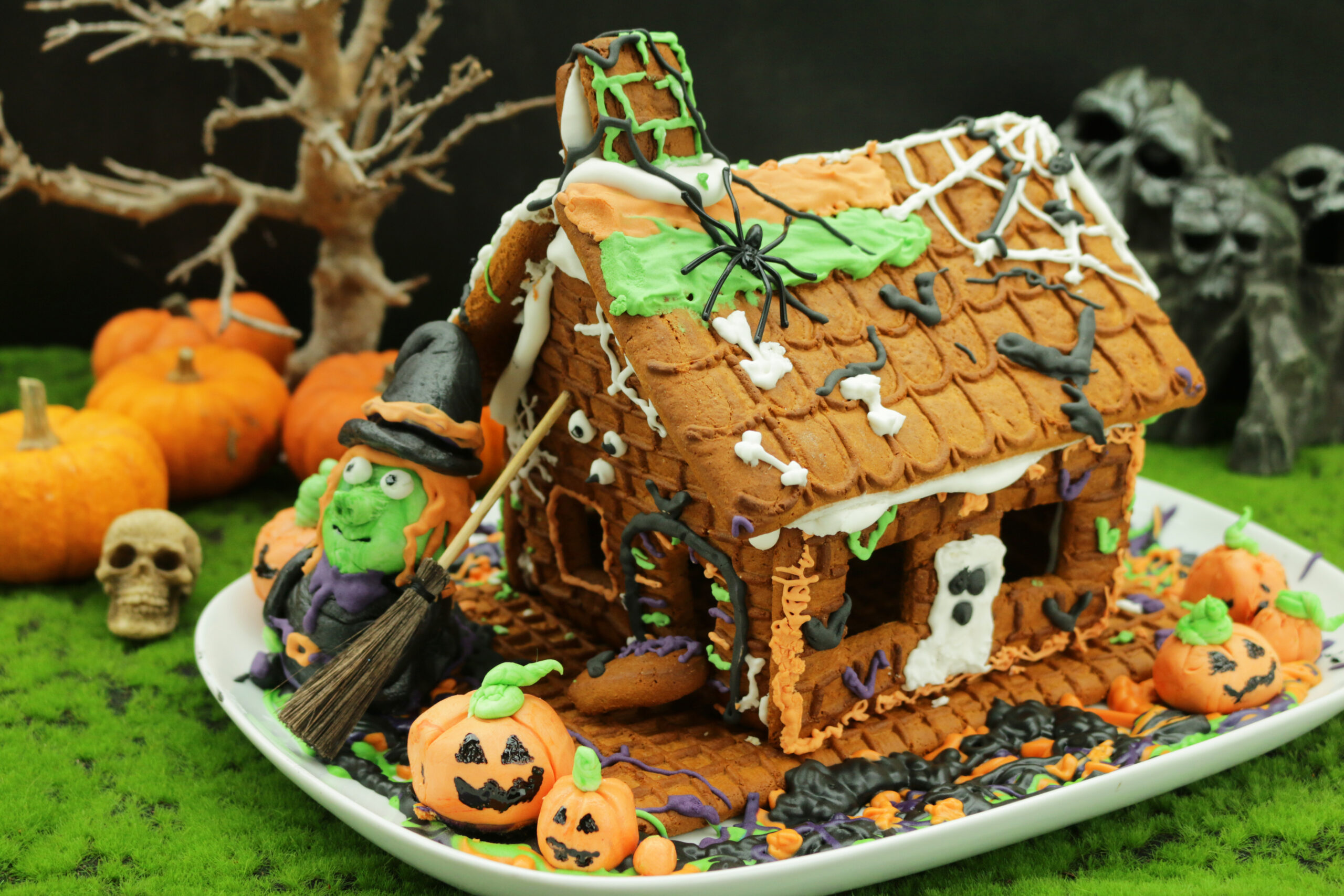 Image of homemade Halloween gingerbread biscuit house decorated with icing and candy sweets on moss worktop, pumpkins and witch figurine