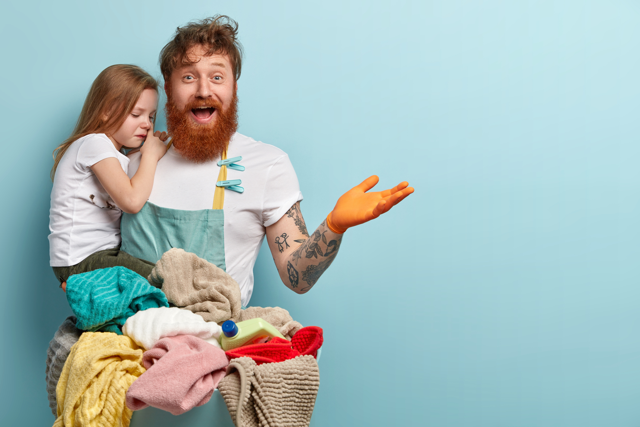 Laundry and household concept. Delighted redhead man with thick beard, raises hand, carries small child with sorrowful expression, isolated over blue background with empty space for text. Busy dad