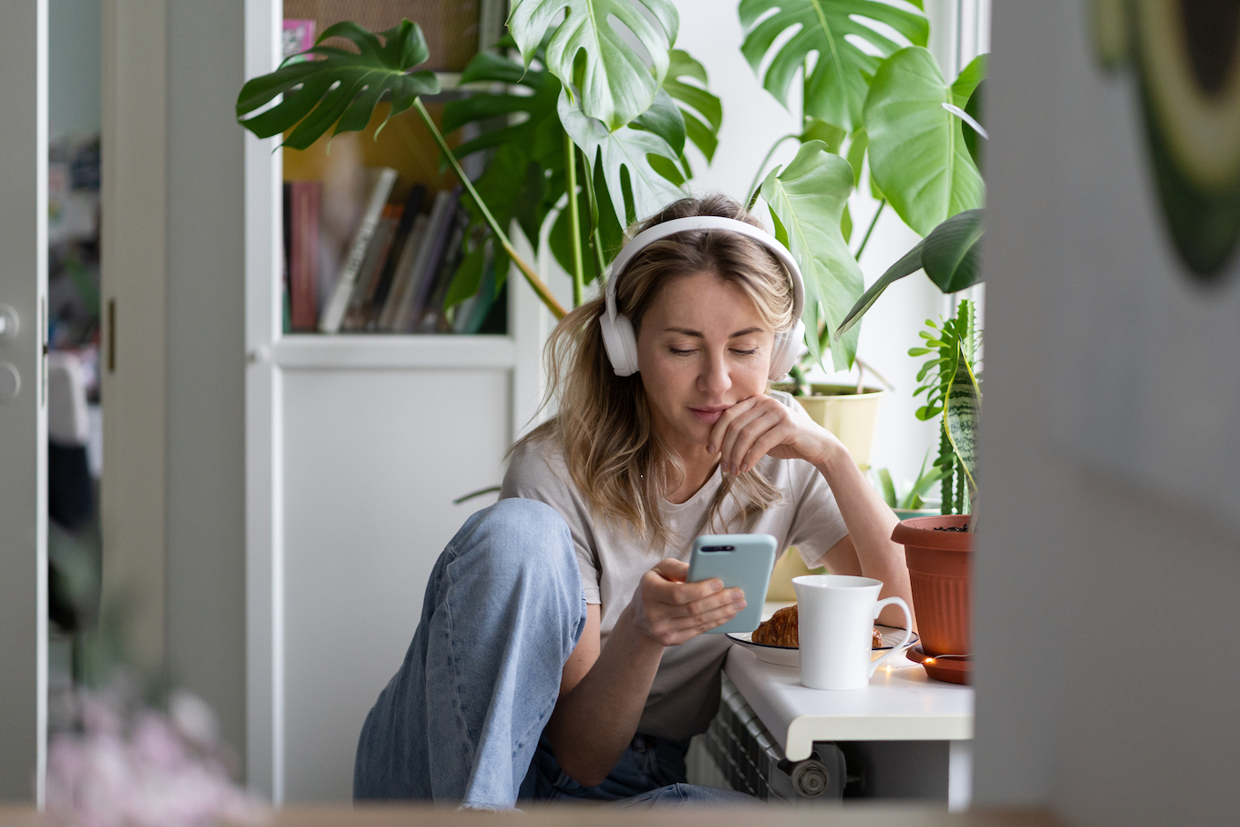 Woman listening to music wear wireless white headphones using mobile smart phone, drinks tea at home