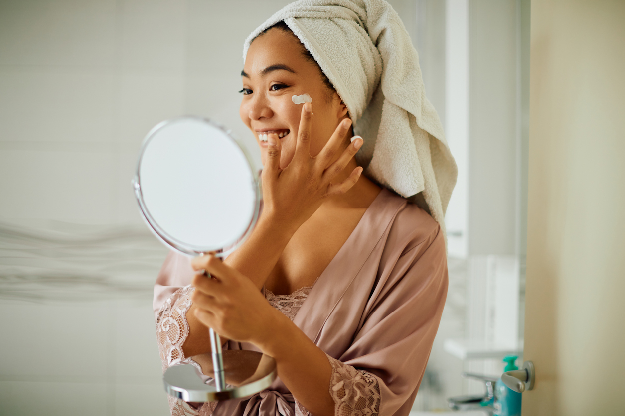 Happy Asian woman applying face cream while looking herself in a mirror in the bathroom.