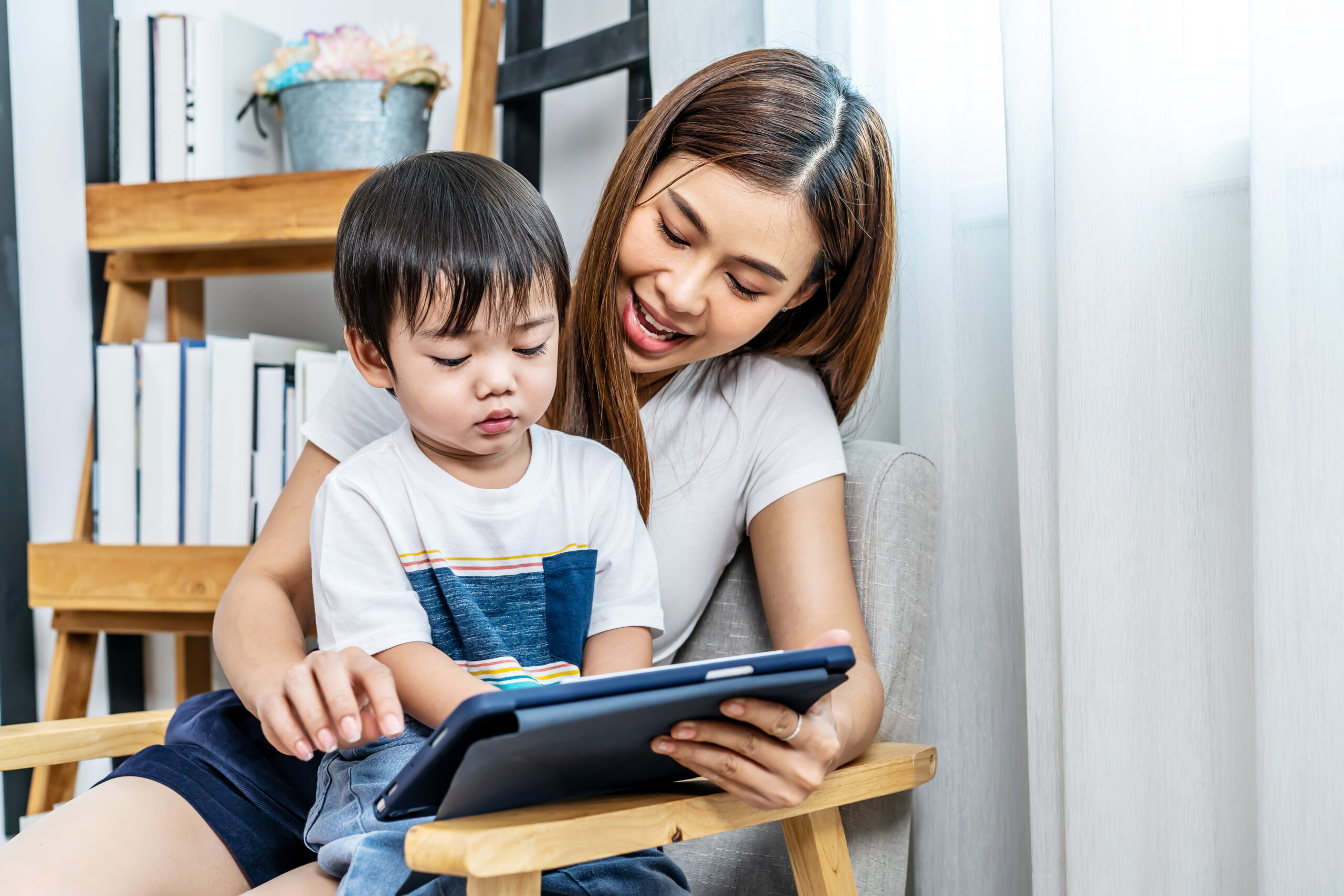 Young asian mother her little adorable son using tablet looking at screen enjoying watching funny social media video doing online shopping relaxing on sofa at home together