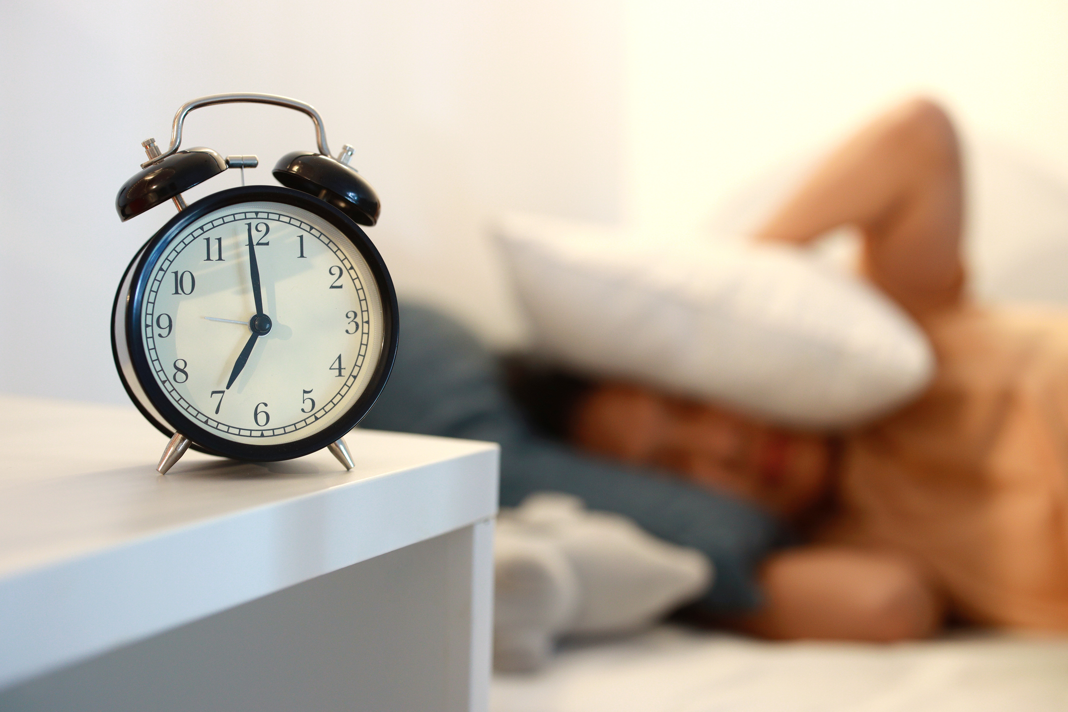 Young asian girl lying in bed and covered by a pillow over her head with alarm clock in early morning
