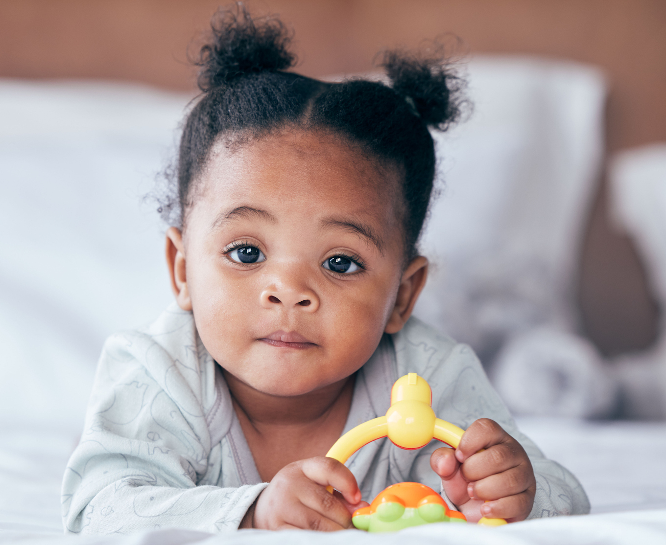 Cute, portrait and baby with a toy on her bed playing and relaxing in her modern nursery. Childhood, playful and African girl infant or newborn playing with plastic object in her bedroom at her home.