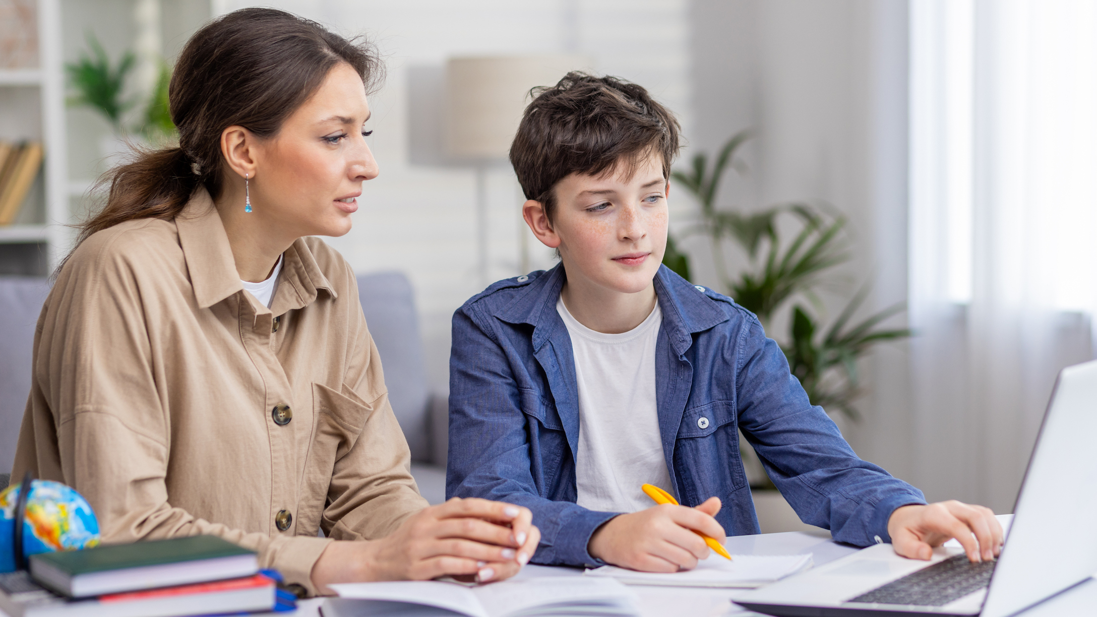 Mother and son studying together sitting at desk at home in living room, boy and woman tutor teaching at home, doing homework together