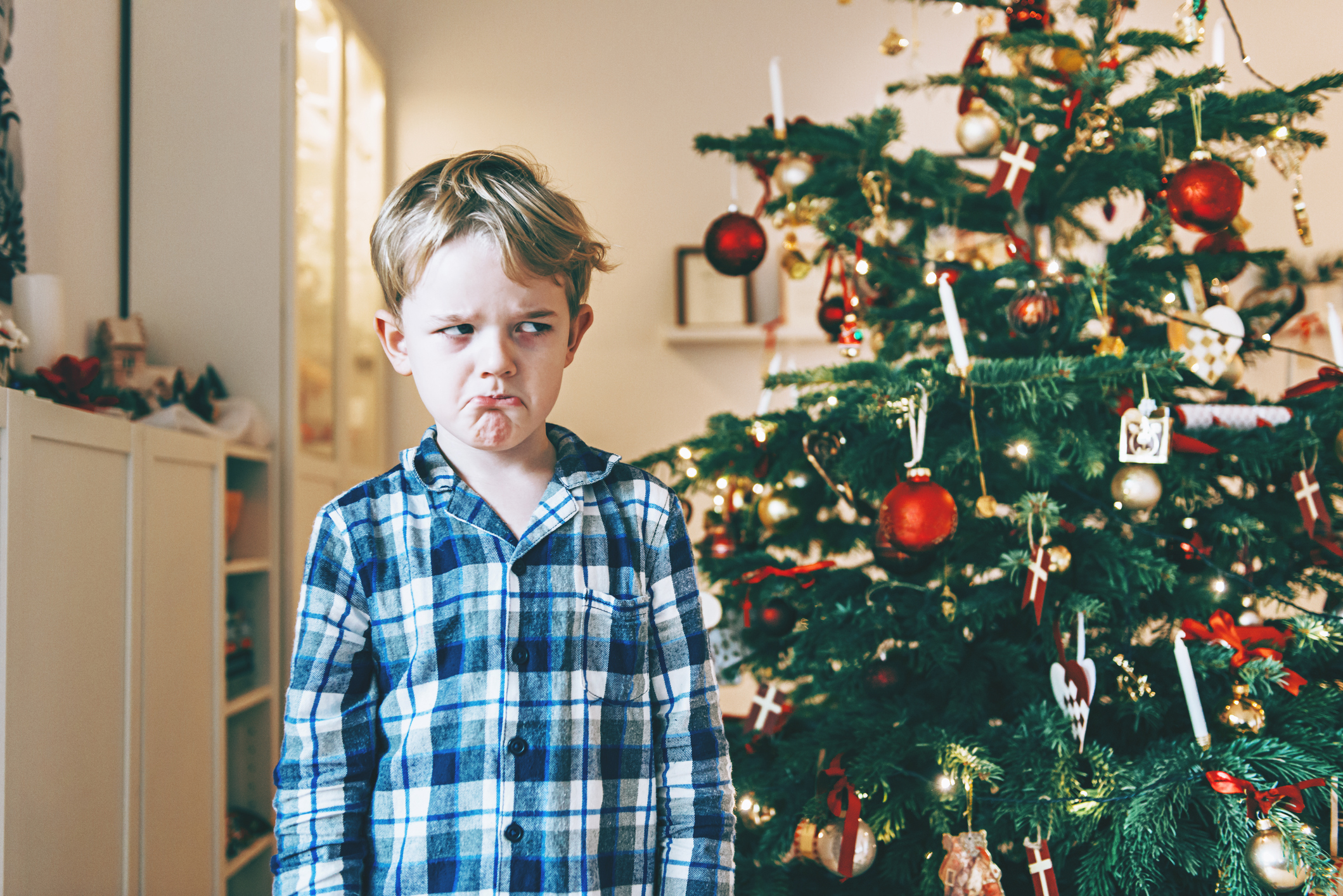 Unhappy boy stands before chistmas tree and makes a face