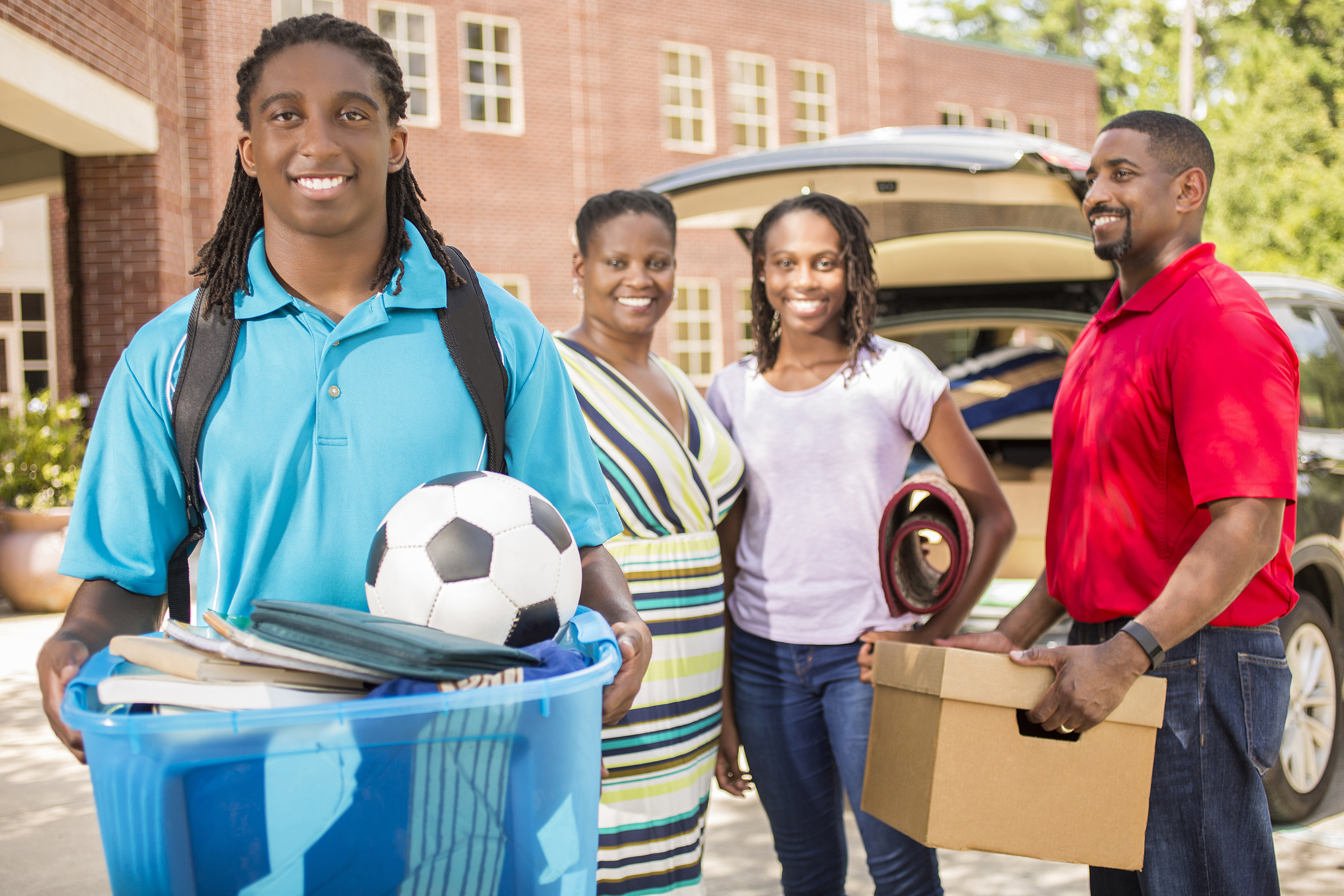 African-descent boy moves into college. Packed car, family. School campus.