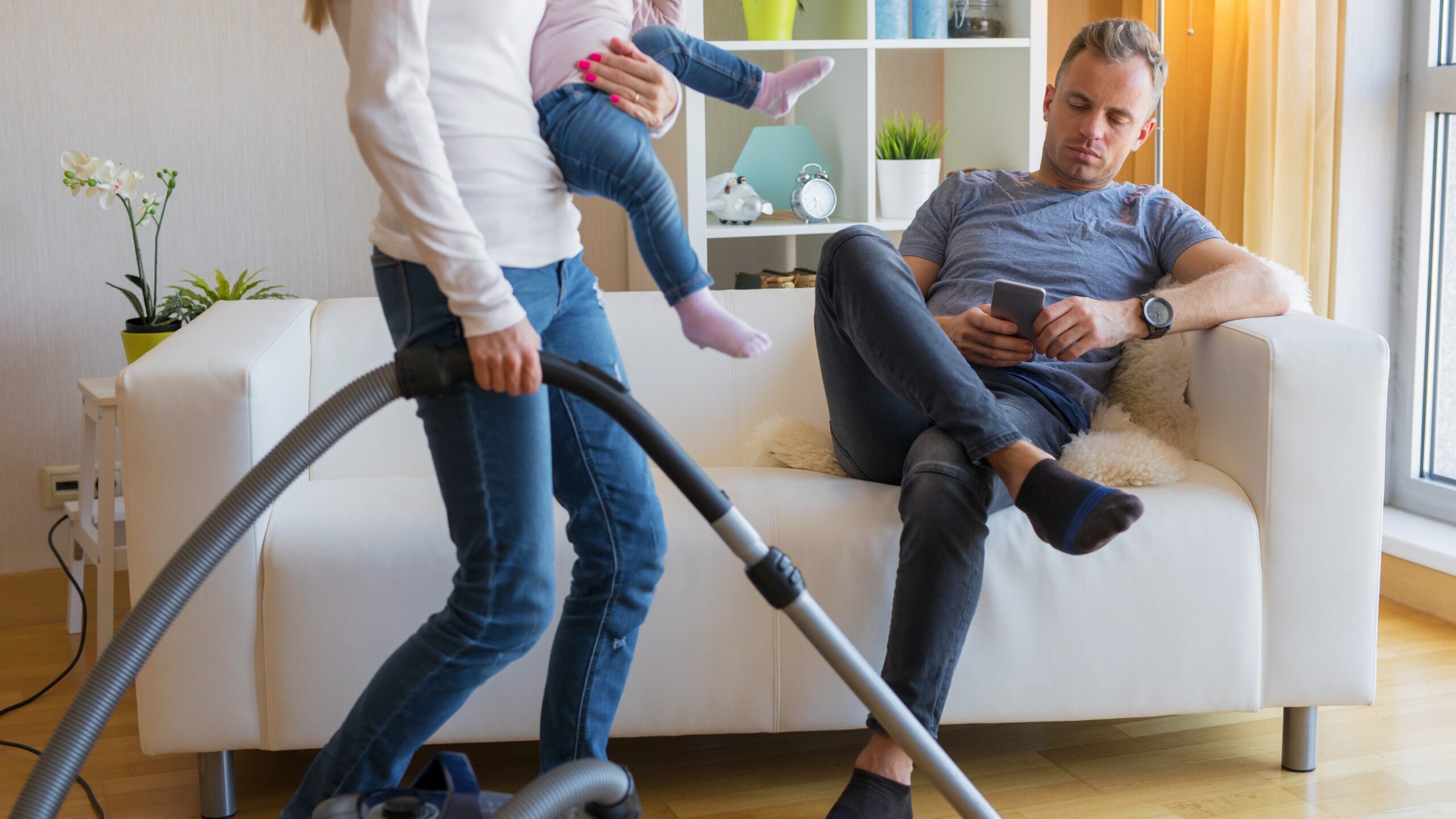 woman doing housework holding baby while husband sits on couch