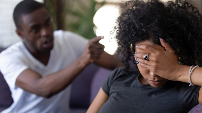 Black man angrily pointing at Black woman who is covering her face with her hand