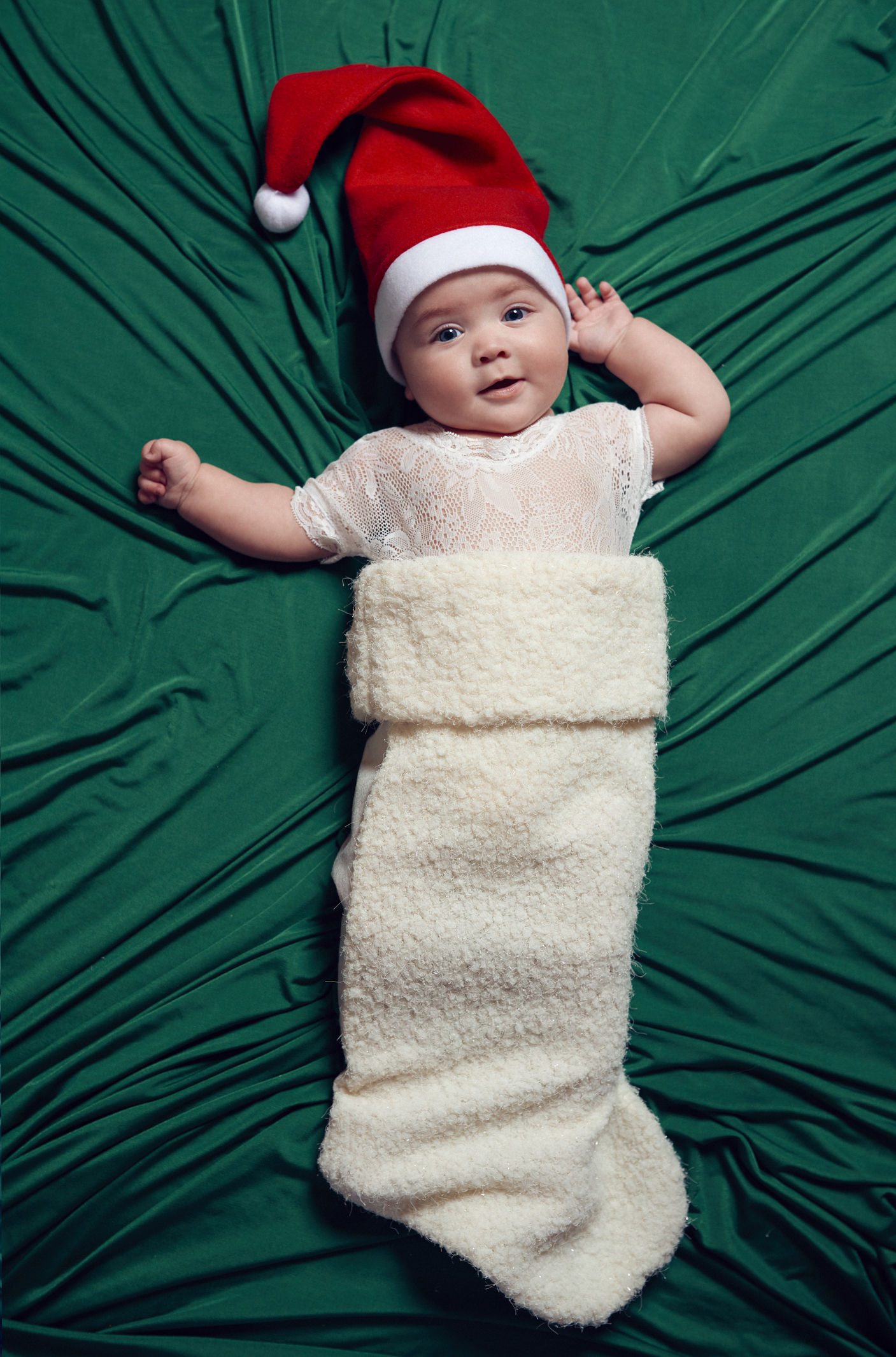 Little baby girl covered in Christmas huge sock and lying on her back on green bed, wearing Christmas hat and looking at the camera happily