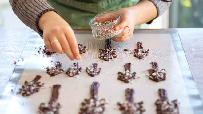 Sustainable baking - vegan candy Christmas Trees. Young woman sprinkling crushed candy cane on dark chocolate trees. Reusable baking mat.