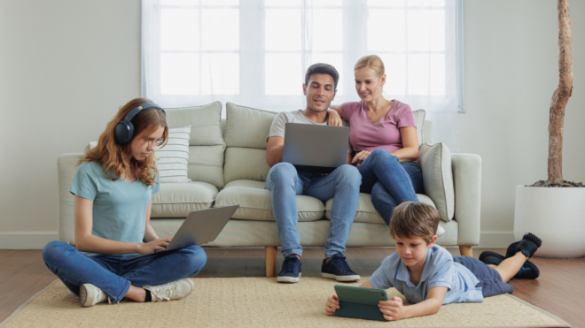 parents sitting on couch sharing a laptop, teen girl sitting on the floor with headphones using laptop, young boy laying on the floor using a tablet
