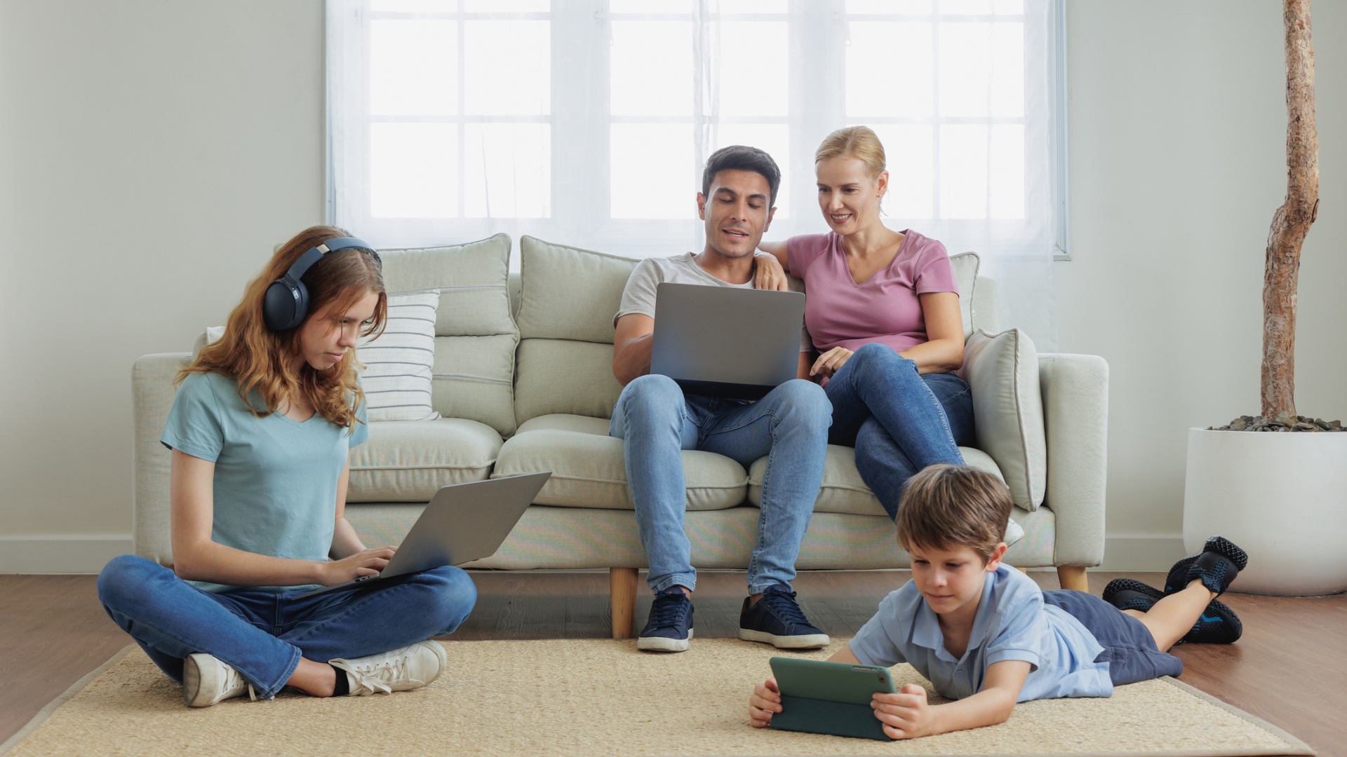 parents sitting on couch sharing a laptop, teen girl sitting on the floor with headphones using laptop, young boy laying on the floor using a tablet