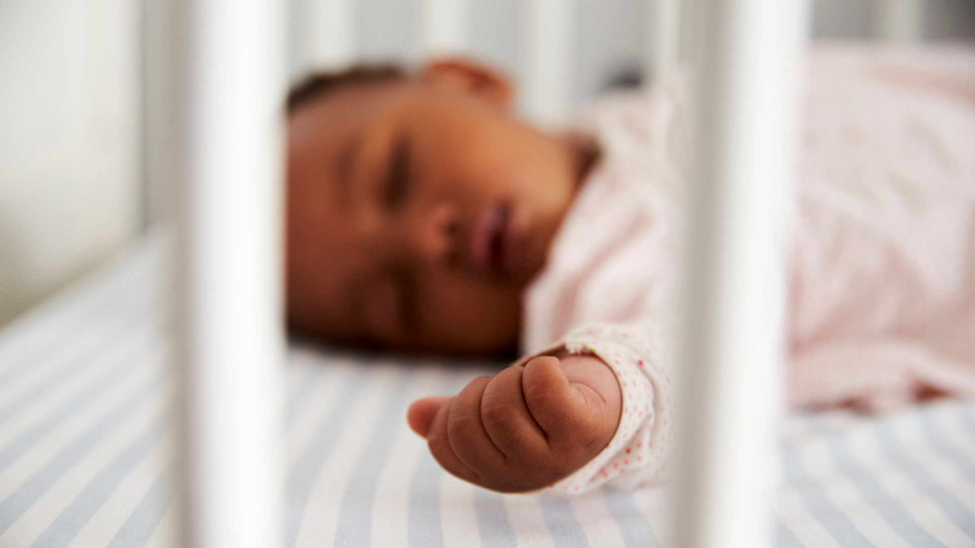 a Black baby laying on their back sleeping through the bars of a crib