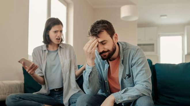 woman holding phone angrily while man holds his head in his hand