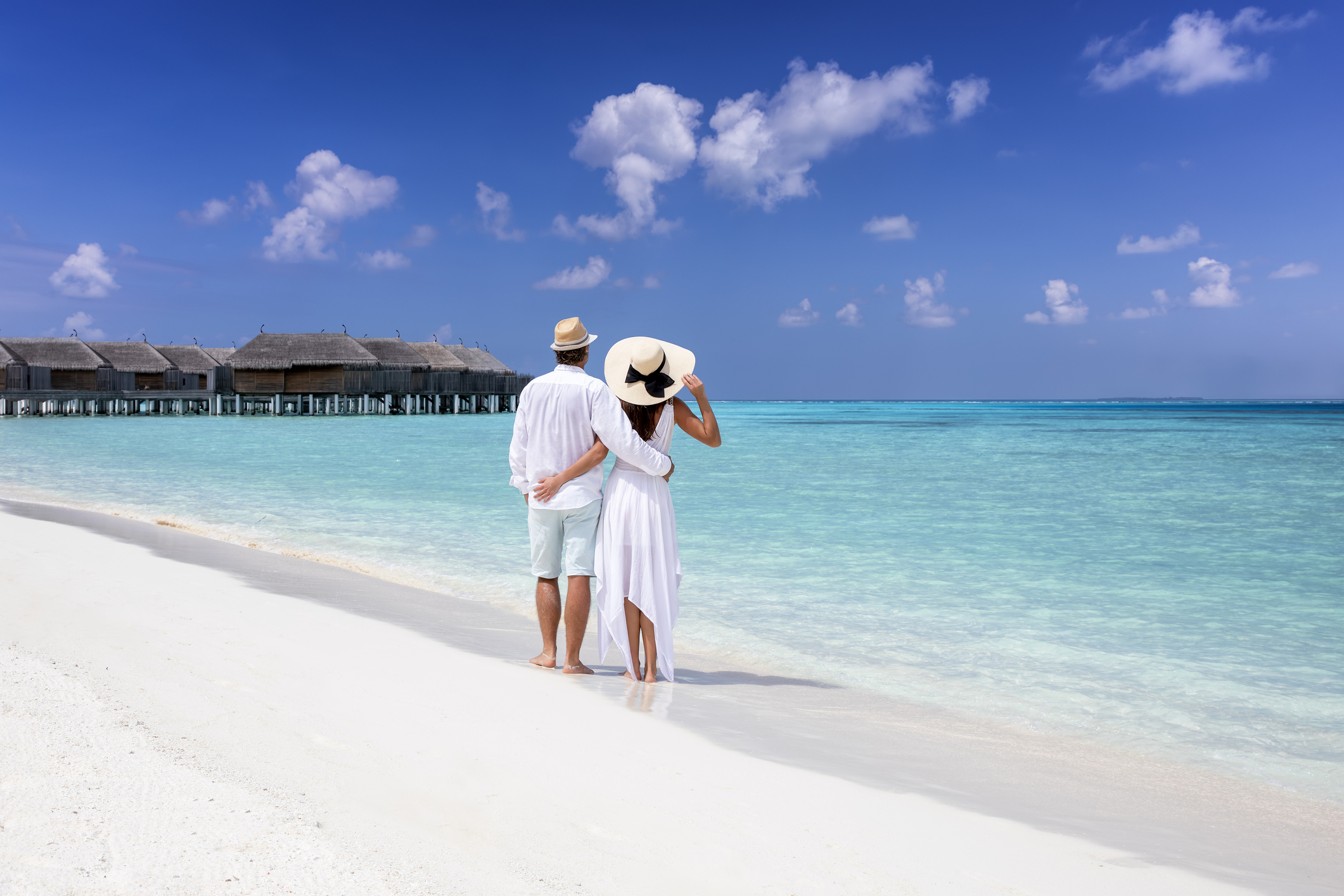 A hugging couple in white summer clothing stands on a tropical beach