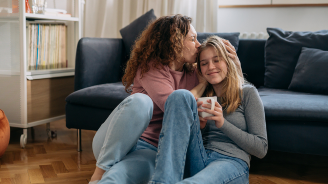 mother with curly hair sitting on floor kissing blonde teen daughter on the head in front of couch