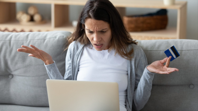 a woman sitting on her couch looking quizzically at a laptop and holding her credit card