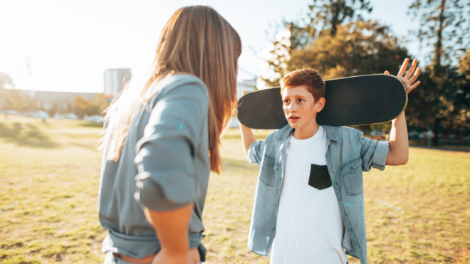 a white mom wearing a gray jacket scolding her son who is holding a skateboard behind his head with his hands open