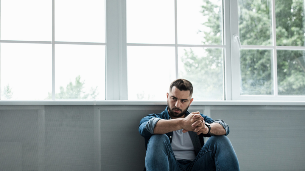white man with beard in denim shirt, white tee and jeans sitting in front of a large window and grey wall