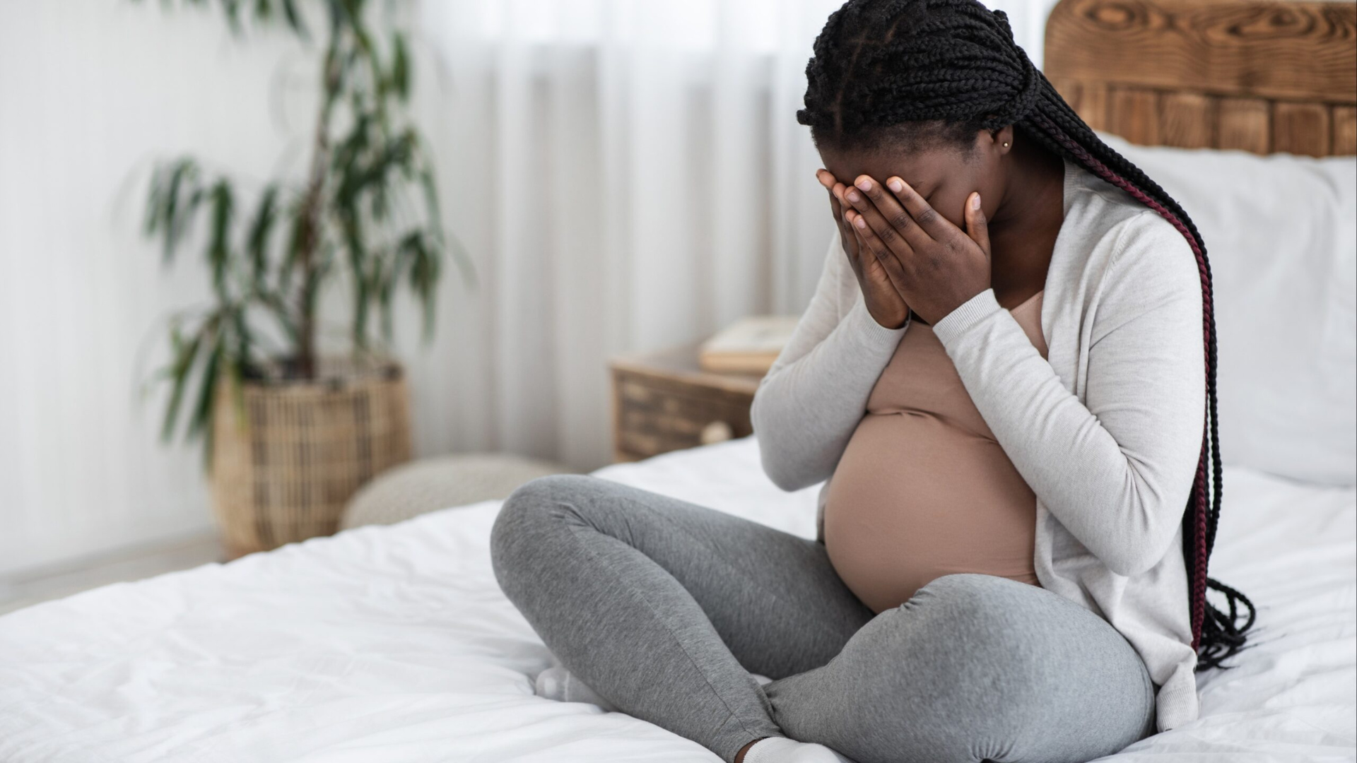 a pregnant Black woman with long braids sitting on a bed with her face in her hands