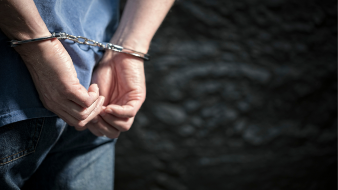 a white person in a blue tee shirt and jeans with their hands behind their back in handcuffs