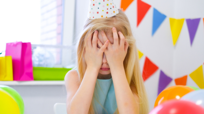 a girl with blonde hair wearing a birthday hat with her face in her hands in front of colorful streamers and presents