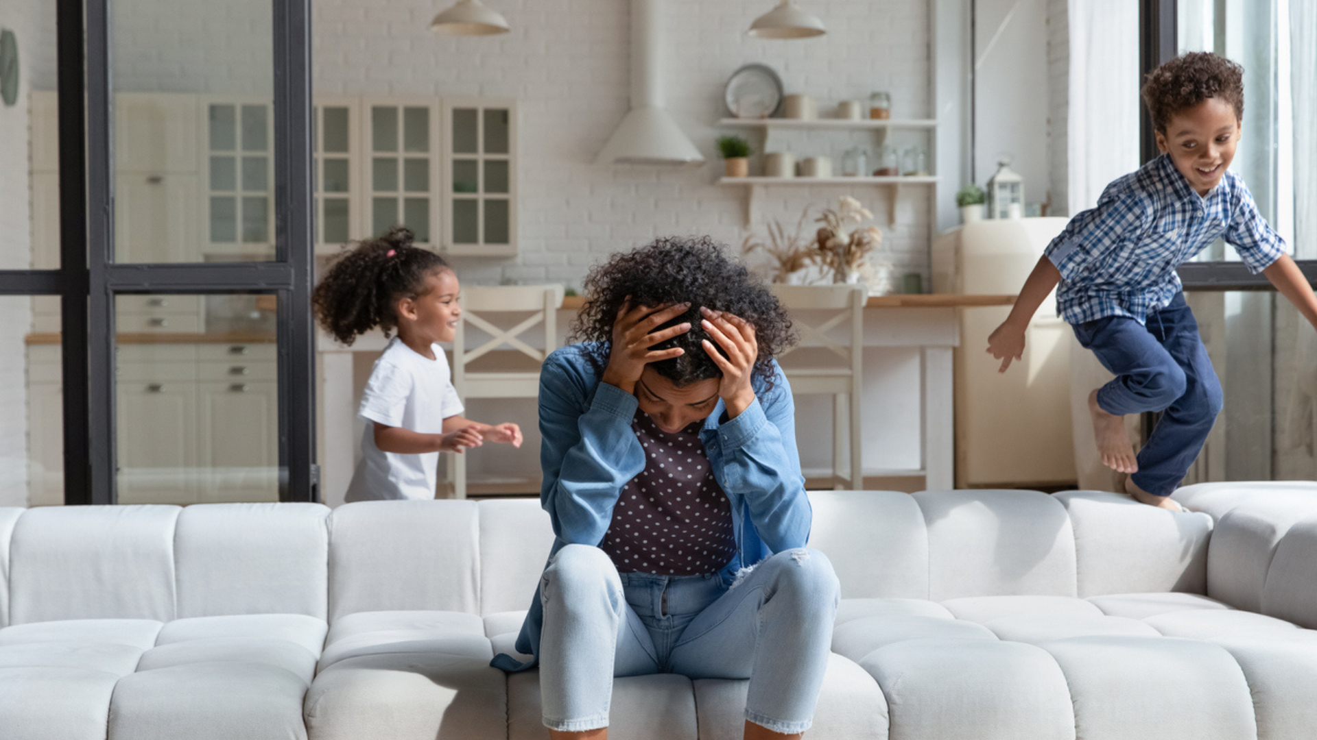 a woman in a denim shirt sitting on the couch with her head in her hands while a young boy and girl run around behind her