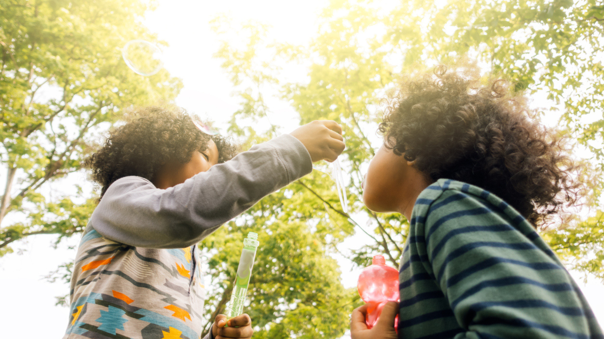 two Black kids with curly hair blowing bubbles with trees above them